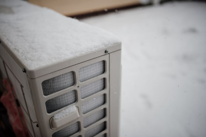 An outdoor air conditioning unit is surrounded by fresh snow, creating a winter wonderland. The equipment is partially obscured as flakes fall softly around it in a quiet, serene e